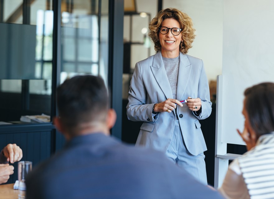 A businesswoman in a light gray suit smiles while leading a discussion with her team in a modern office setting.