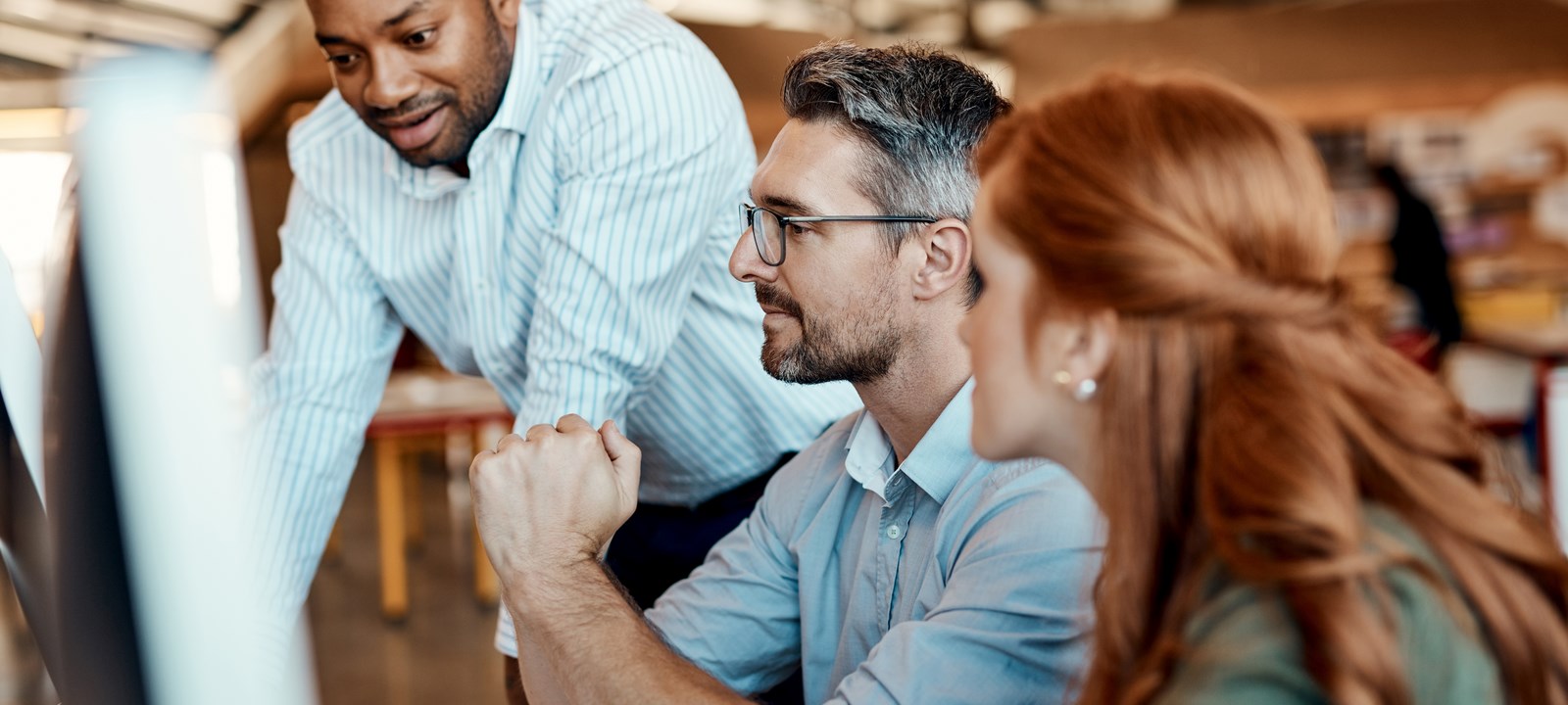 A diverse group of three colleagues collaborating at an office desk, with one person standing and two seated, focusing on a computer screen while discussing ideas.