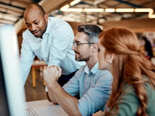 A diverse group of three colleagues collaborating at an office desk, with one person standing and two seated, focusing on a computer screen while discussing ideas.