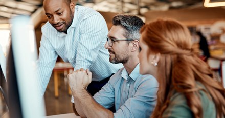 A diverse group of three colleagues collaborating at an office desk, with one person standing and two seated, focusing on a computer screen while discussing ideas.