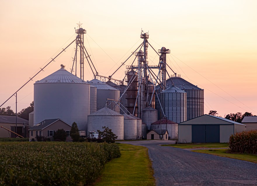 A large industrial farm with several metal grain silos and granary elevators, surrounded by fields and buildings, set against a soft sunset sky.