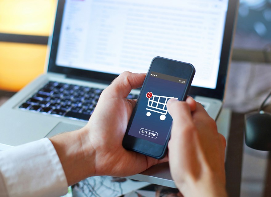 A person holding a smartphone with an online shopping cart interface, showing two notifications and a 'Buy Now' button, with a laptop in the background, representing e-commerce or online shopping.
