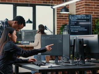 A diverse team of professionals working in an office setting, collaborating on data tasks at multiple computer workstations, with code displayed on monitors and a modern workspace environment.