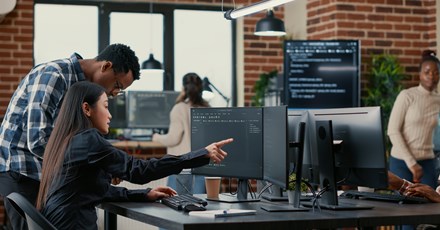 A diverse team of professionals working in an office setting, collaborating on data tasks at multiple computer workstations, with code displayed on monitors and a modern workspace environment.
