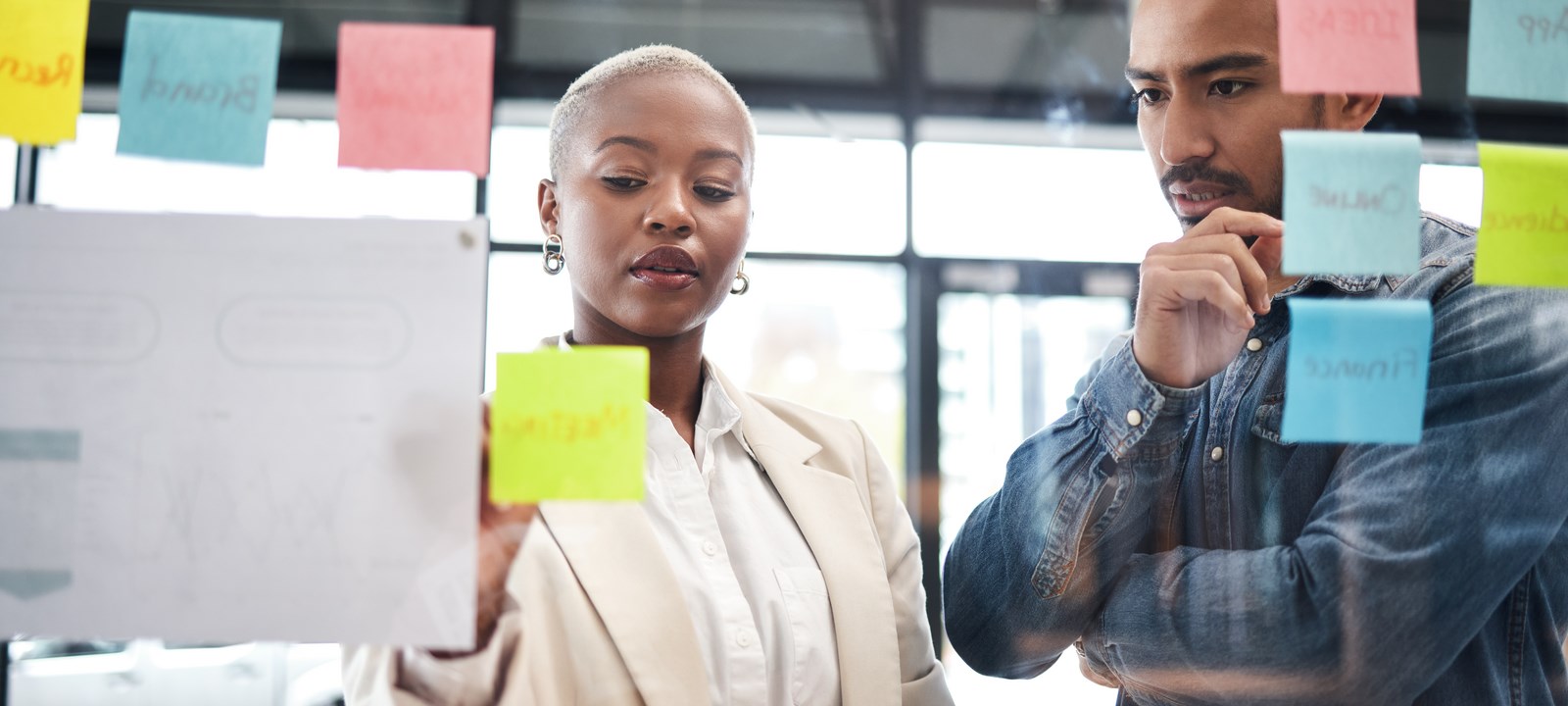 Two professionals are engaged in a collaborative brainstorming session, using a glass wall covered with colorful sticky notes. The woman, with short blonde hair and wearing a beige blazer, is placing a yellow sticky note on the wall, while the man next to her, dressed in a denim shirt, observes thoughtfully with his hand on his chin.