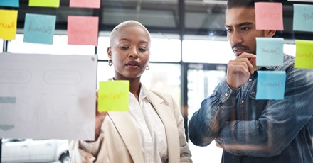 Two professionals are engaged in a collaborative brainstorming session, using a glass wall covered with colorful sticky notes. The woman, with short blonde hair and wearing a beige blazer, is placing a yellow sticky note on the wall, while the man next to her, dressed in a denim shirt, observes thoughtfully with his hand on his chin.