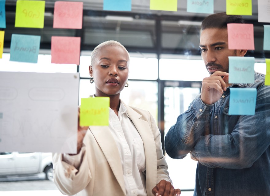 Two professionals are engaged in a collaborative brainstorming session, using a glass wall covered with colorful sticky notes. The woman, with short blonde hair and wearing a beige blazer, is placing a yellow sticky note on the wall, while the man next to her, dressed in a denim shirt, observes thoughtfully with his hand on his chin.