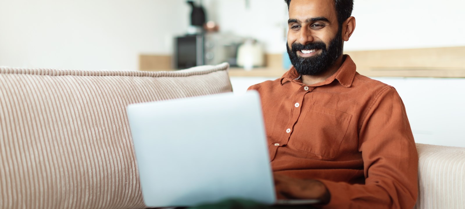 A man sitting on a couch smiles while shopping online using his laptop in a comfortable, modern home setting.