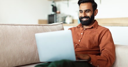 A man sitting on a couch smiles while shopping online using his laptop in a comfortable, modern home setting.