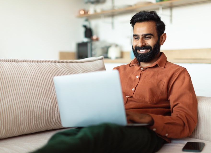 A man sitting on a couch smiles while shopping online using his laptop in a comfortable, modern home setting.