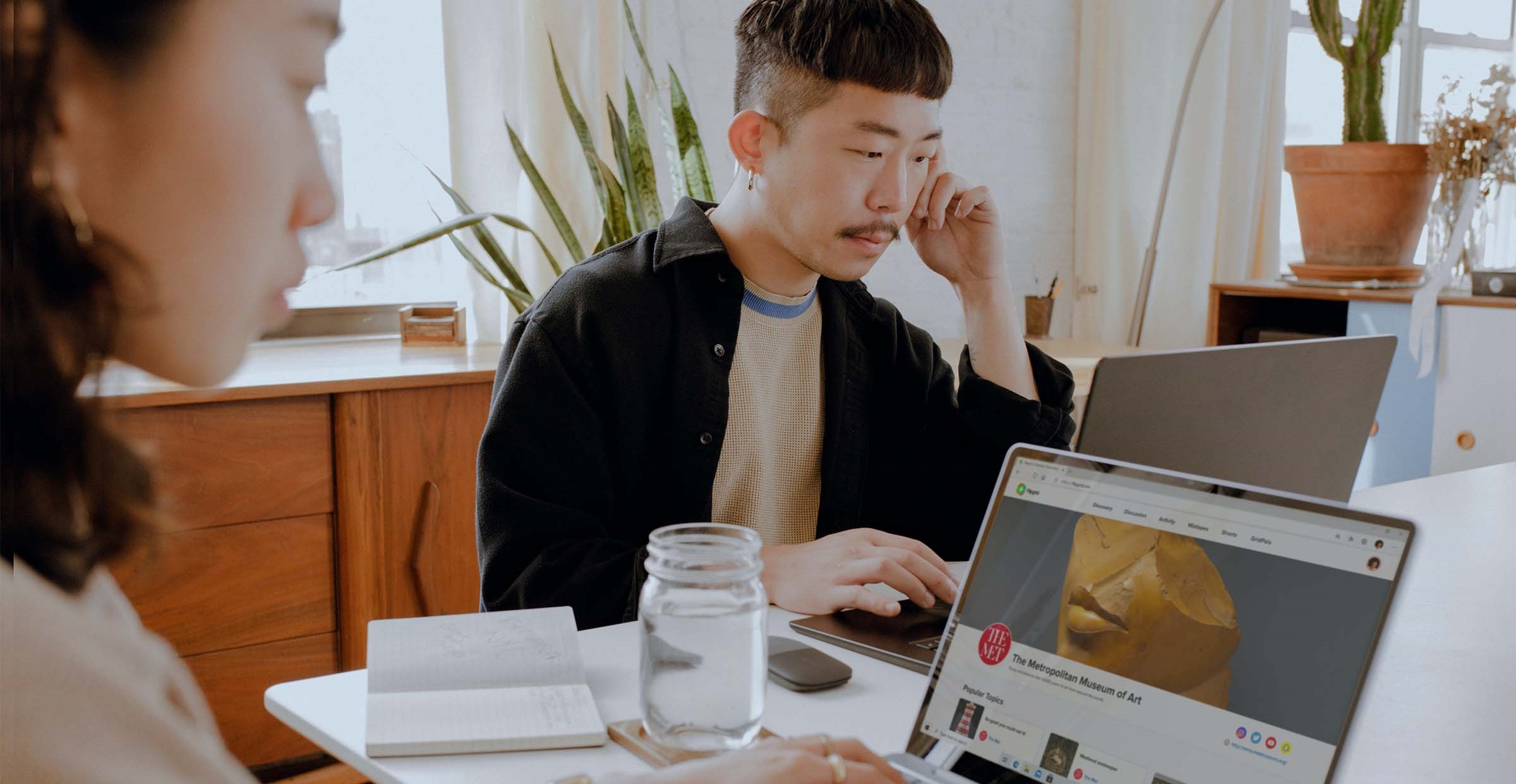 Two people of Asian descent sit at a table looking at their separate computers but working together