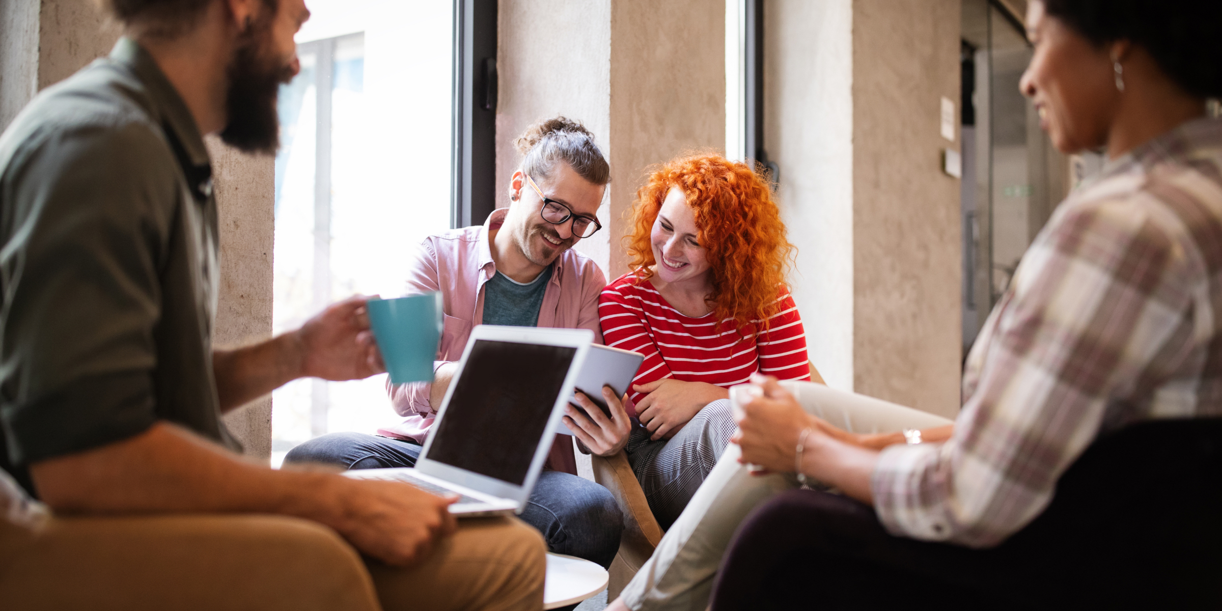 4 people in an office talking. The main focus is on a man with glasses and brown hair and a woman with red hair, smiling at a laptop